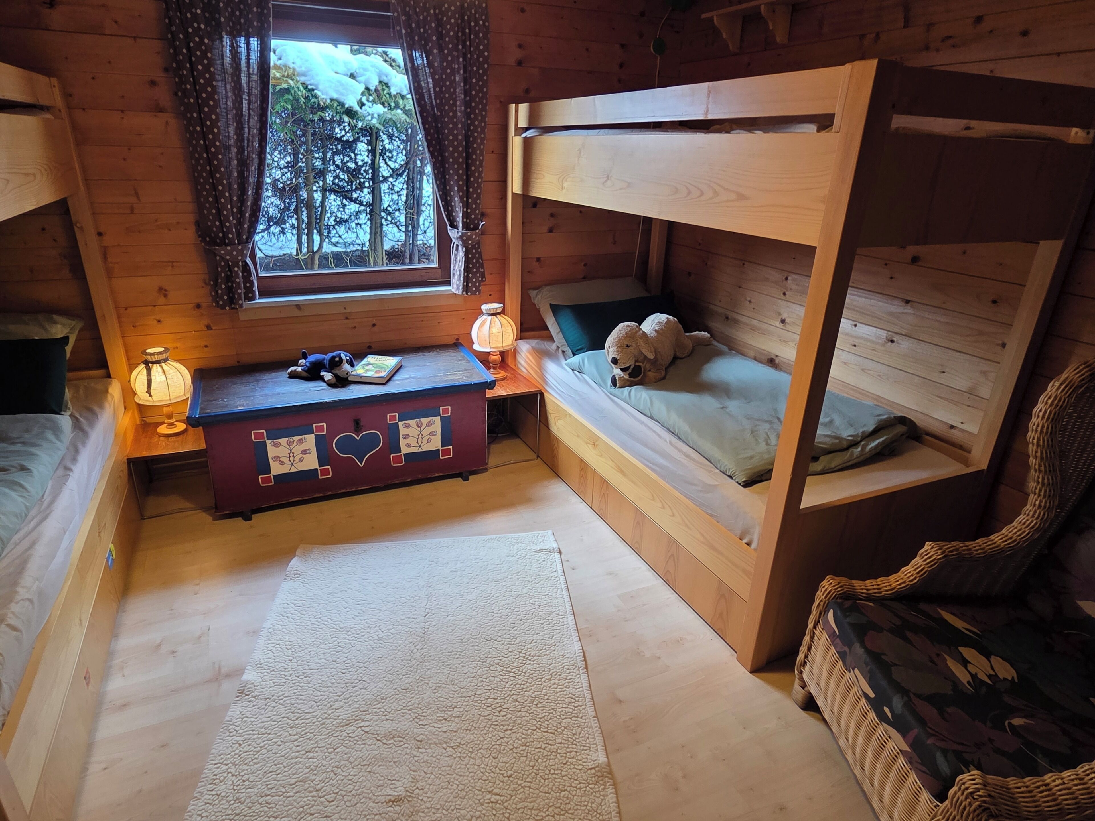 A cozy cabin room with a bunk bed, a small desk with a lamp, a teddy bear, and a view of trees through a window.