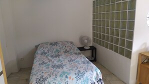 Una habitación pequeña y sencilla, con cama, ventilador y pared de azulejos.