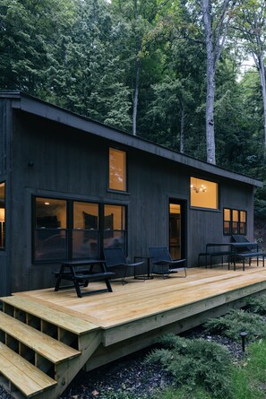 A modern wooden cabin with a deck, large windows, and a view of the forest.