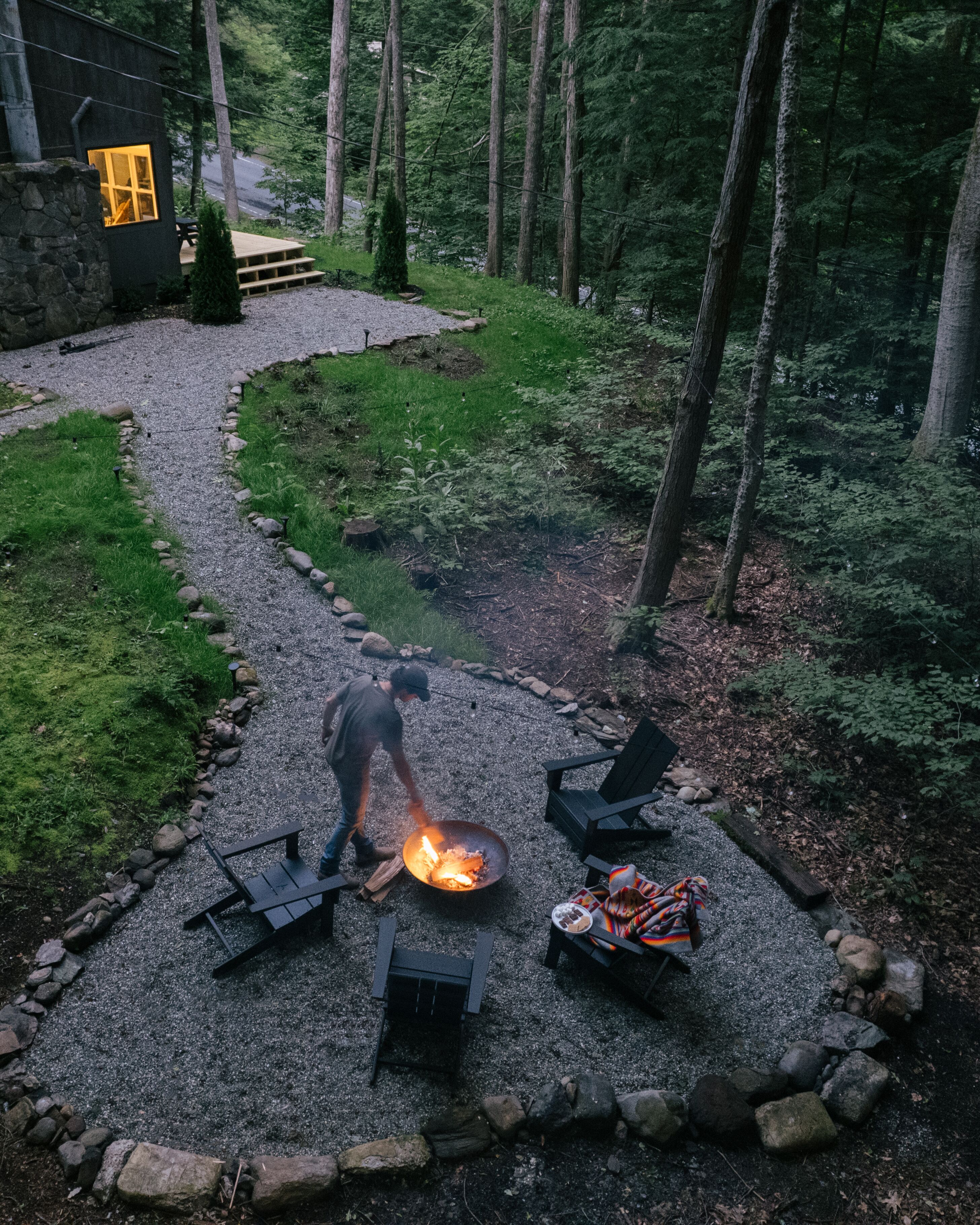 A stone pathway leading to a rustic outdoor seating area with a fire pit, surrounded by a forested landscape.