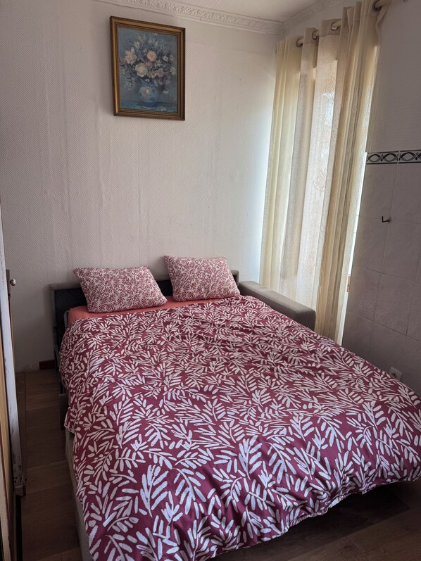 A bedroom with a bed, two pillows, a painting on the wall, and a window with curtains. - At DAGHSEN (Nice)