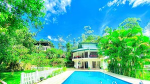 A pool area with a two-story building, surrounded by lush greenery and a white fence. - Blue Mountain Resort Bandarawela  (Bandarawela)