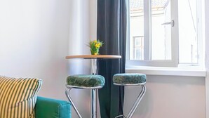 Un rincón de una habitación con una ventana, una mesita con un florero y dos taburetes de bar.