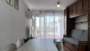Un espace repas comprenant une table et des chaises, un canapé, un luminaire suspendu et une vue par une fenêtre ornée de rideaux légers.