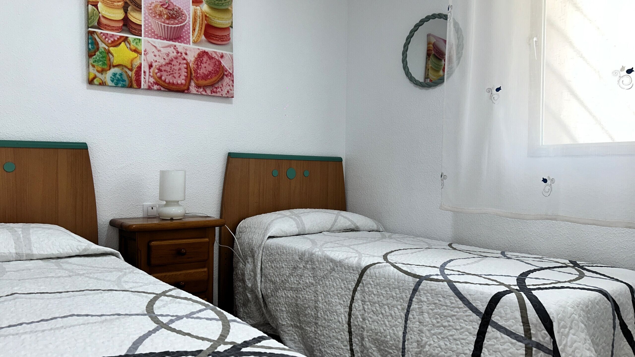A bedroom with two beds, a nightstand with a lamp, a wall clock, and a window with curtains.