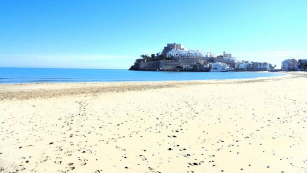 Une plage de sable où l’on peut voir des empreintes de pas menant à une structure fortifiée, avec des bâtiments blancs et un château au sommet, d’où l’on a une vue sur la mer.