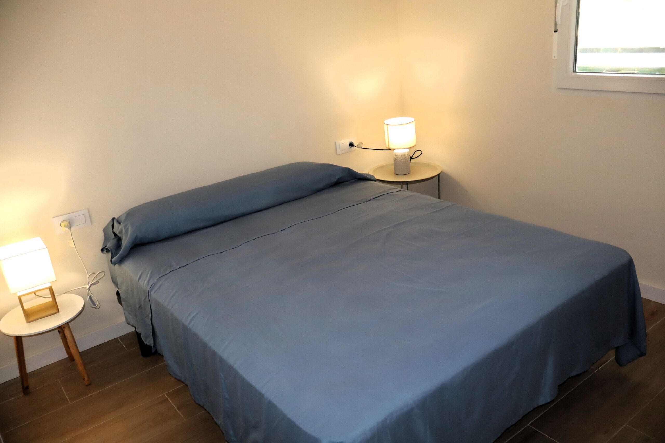A single bed with a blue bedspread, a bedside table with a lamp, and a power outlet on the wall.