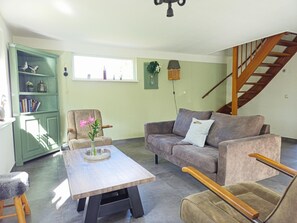 Een woonkamer met een bank, fauteuil en een tafel met een vaas. Een trap en een groene kast met decoratieve objecten.