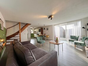Een moderne woonkamer met een bruine leren bank, groene stoelen, een houten eettafel en een keuken op de achtergrond.