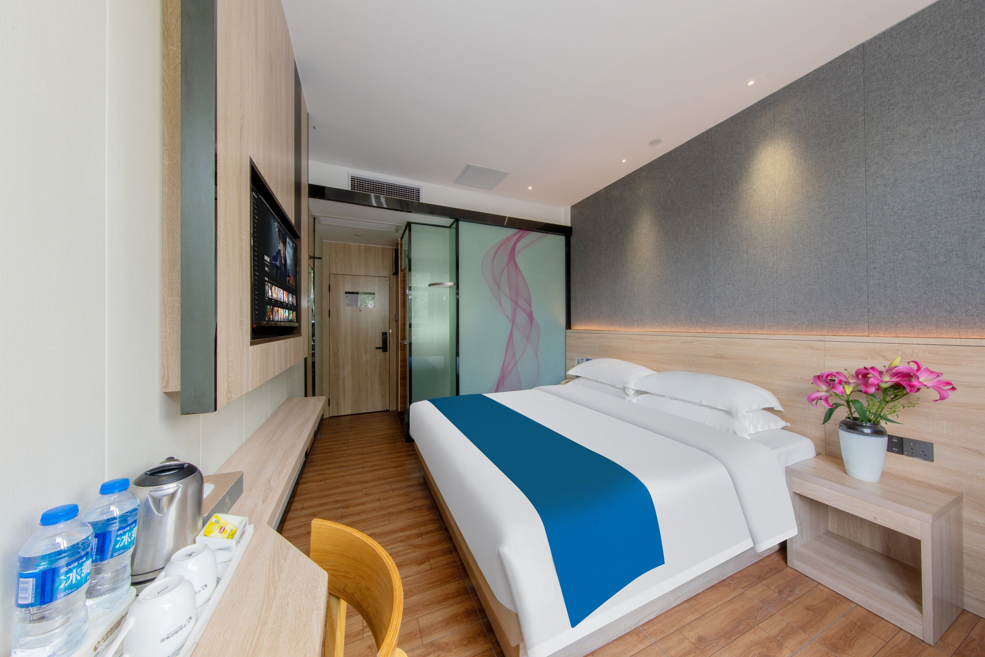 Una habitación de hotel moderna con una cama grande, una mesita de noche y un pequeño escritorio con una silla. Hay un televisor de pared y una planta en maceta sobre la mesita de noche.