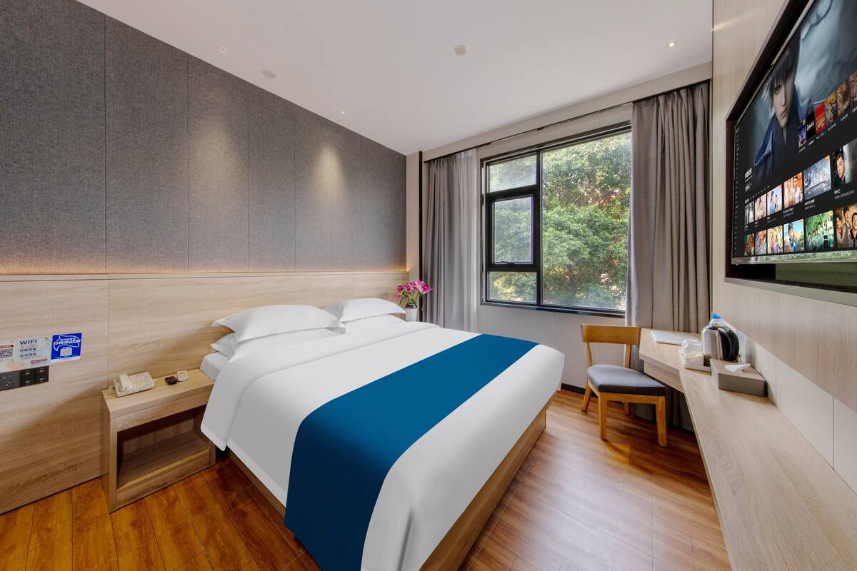 Una habitación de hotel moderna con una cama grande, un escritorio, una silla, un televisor y una ventana con vistas a árboles.