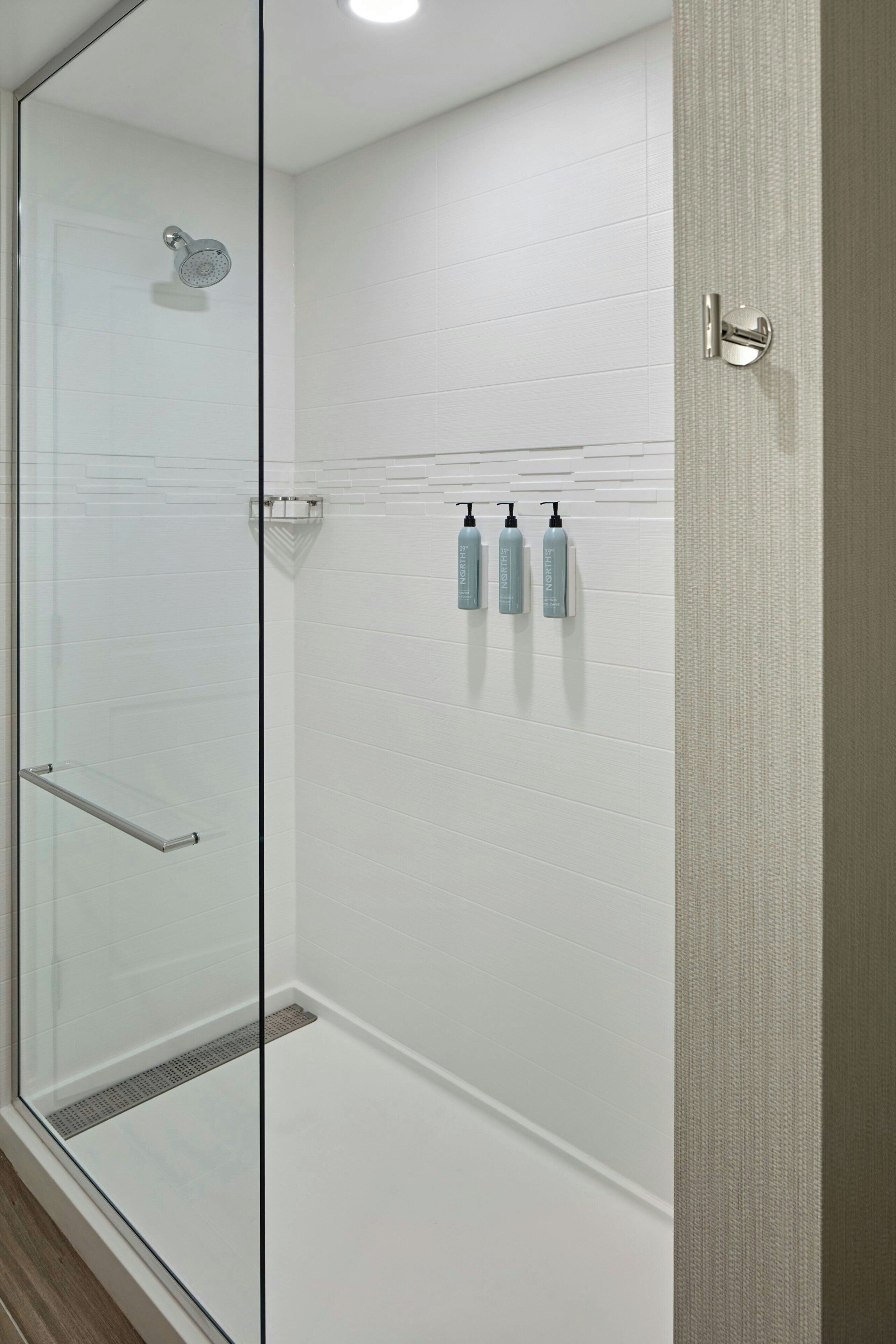Ducha con mampara de vidrio, pared de azulejos blancos, puerta de vidrio y un perchero con botellas.