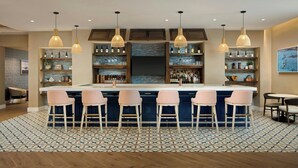 Eine Bar mit einer zentralen Insel, Barhocker, Pendelleuchten, Regalen mit Flaschen und einem gemusterten Boden.