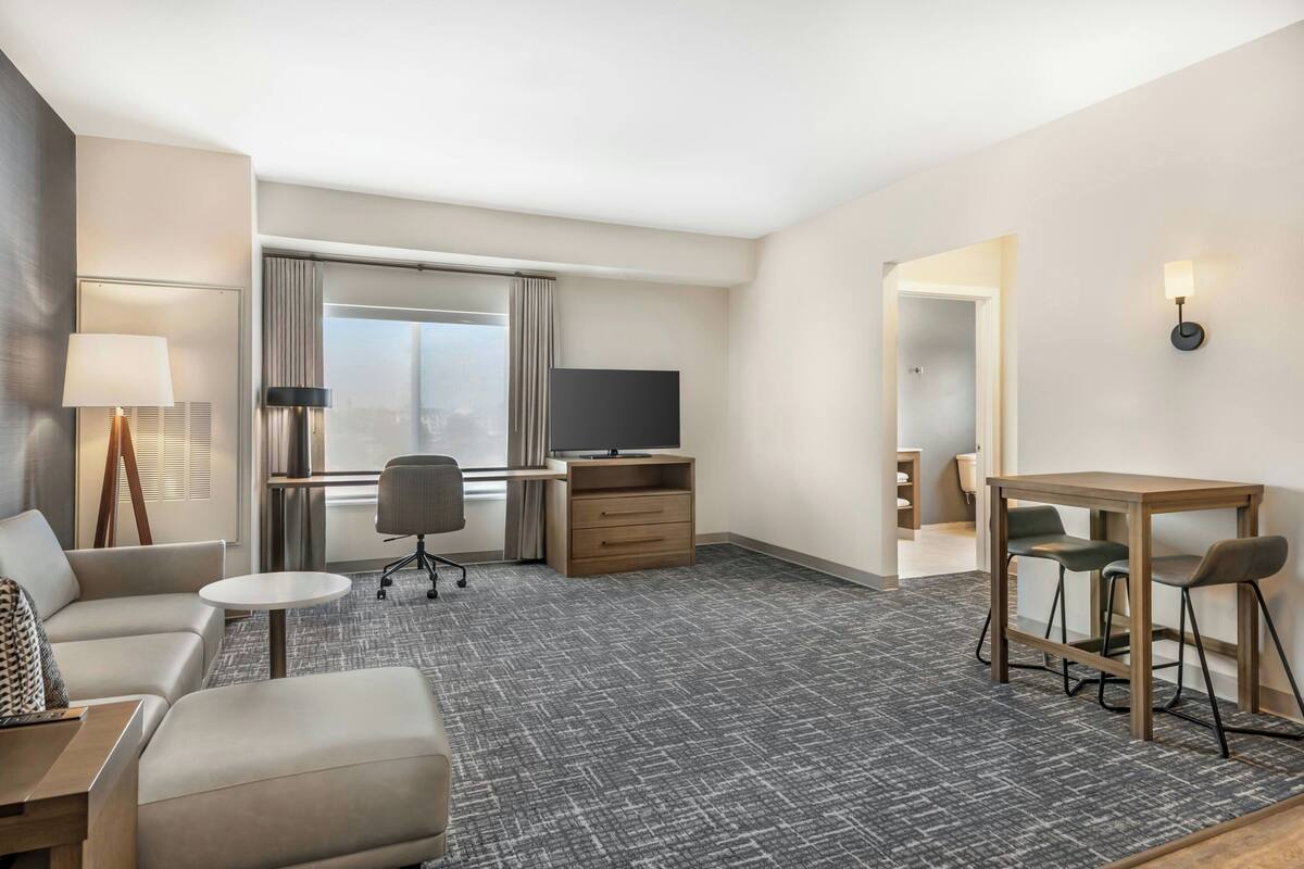 A hotel room with a sofa, a small round table, a desk with a chair, a TV, and a large window with curtains.