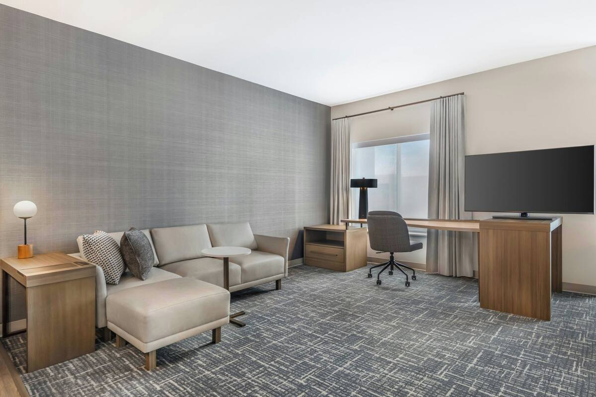 A hotel room with a sofa, desk, chair, TV, lamp, and a large window with curtains.