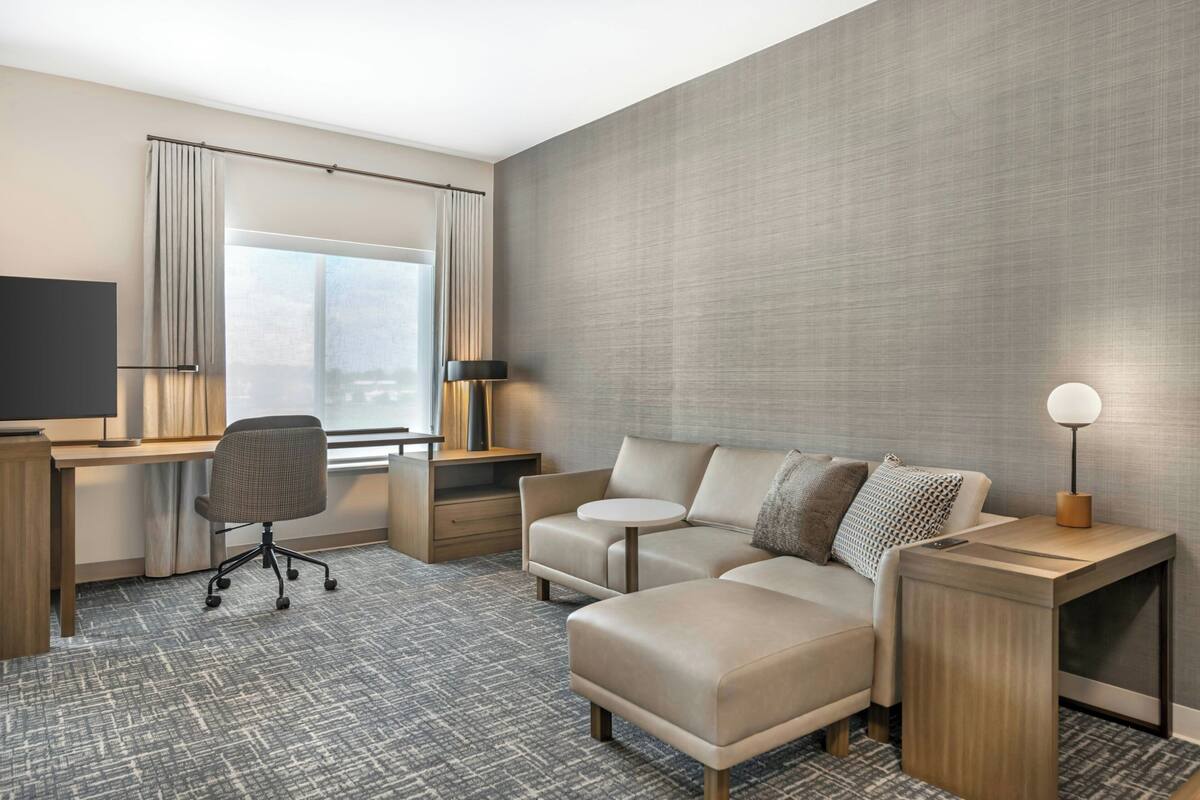 A hotel room with a desk, office chair, sofa, and a bedside table with a lamp.