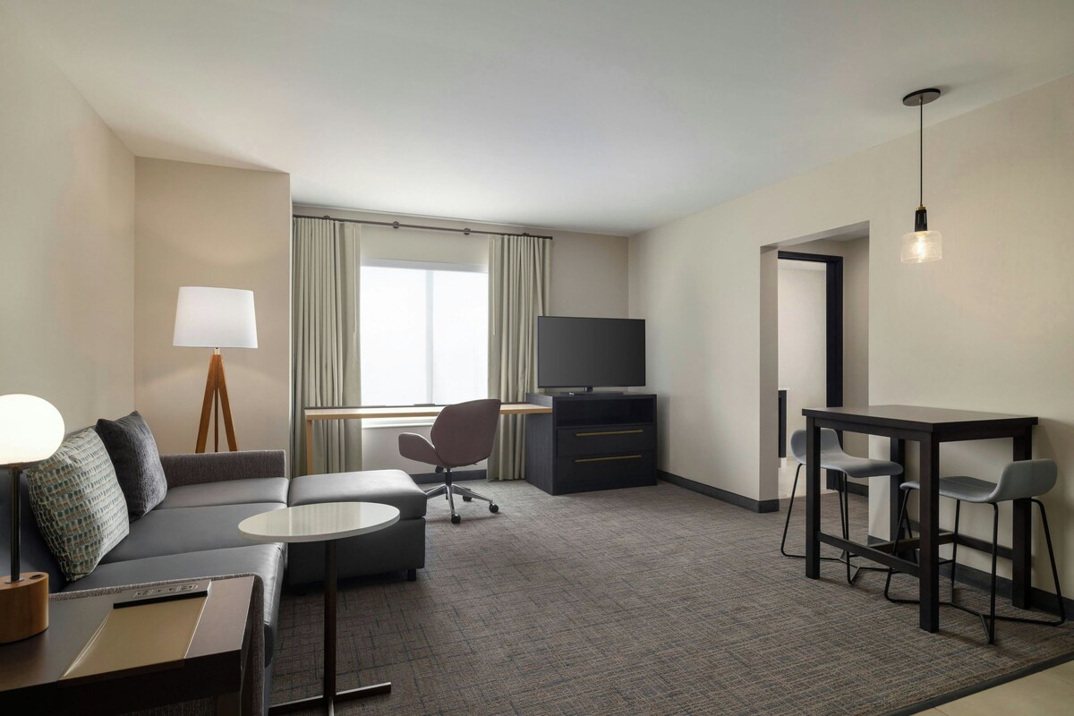 A modern hotel room with a sofa, a desk, a television, and a dining area.