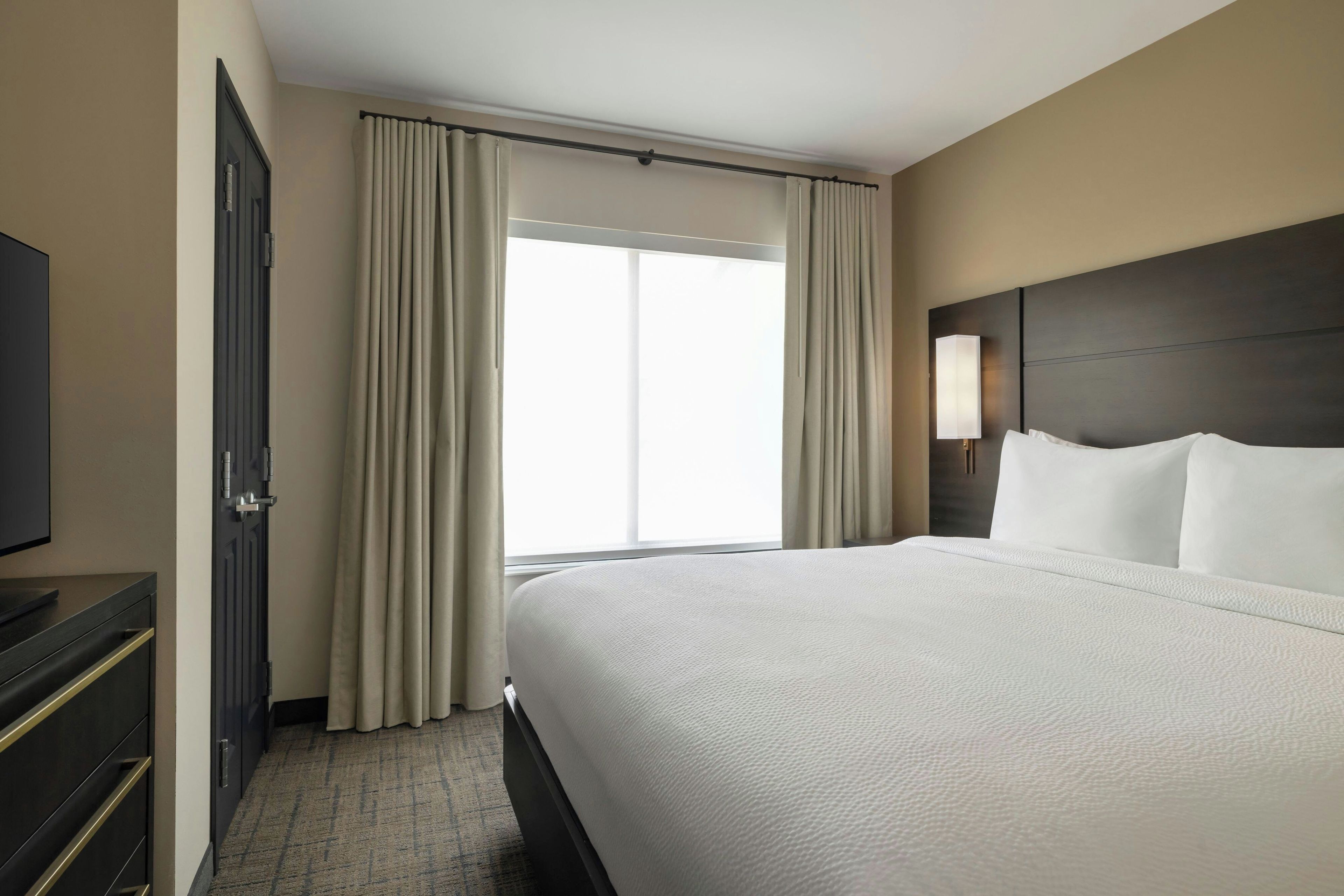 Ein Hotelzimmer mit einem großen Bett, einem Fernseher auf einer Kommode und einem Fenster mit Vorhängen.