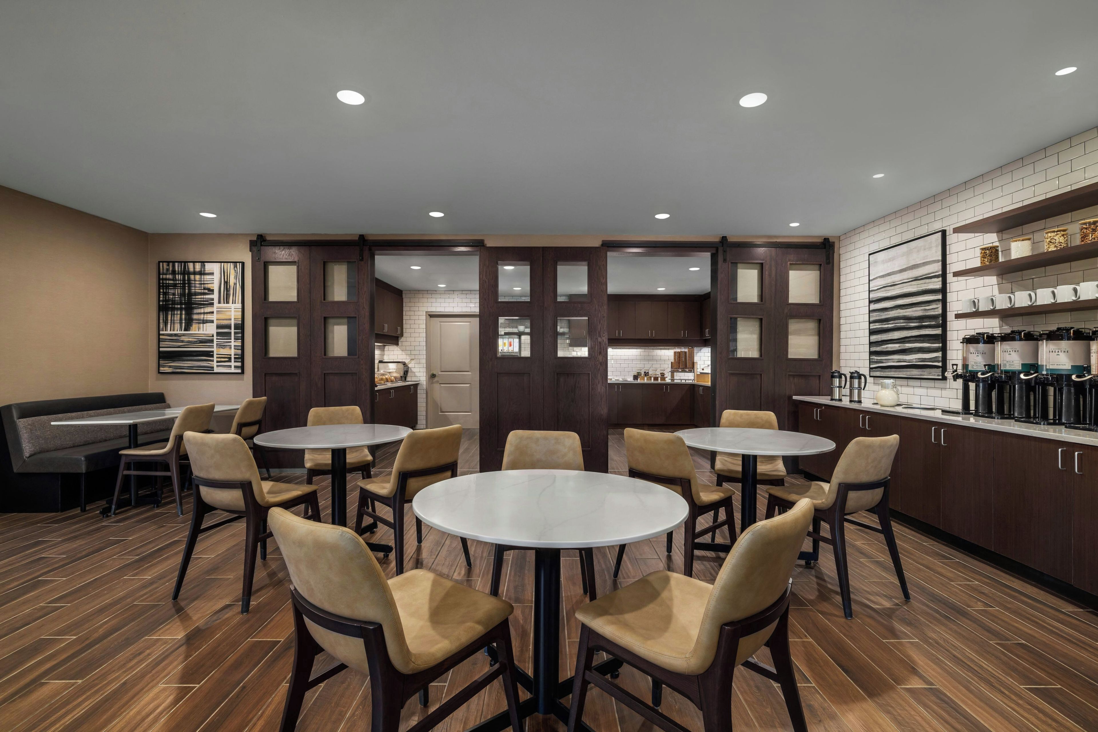 Un interior moderno con mesas y sillas redondas, una zona de bar y una cocina visible a través de un vidrio.