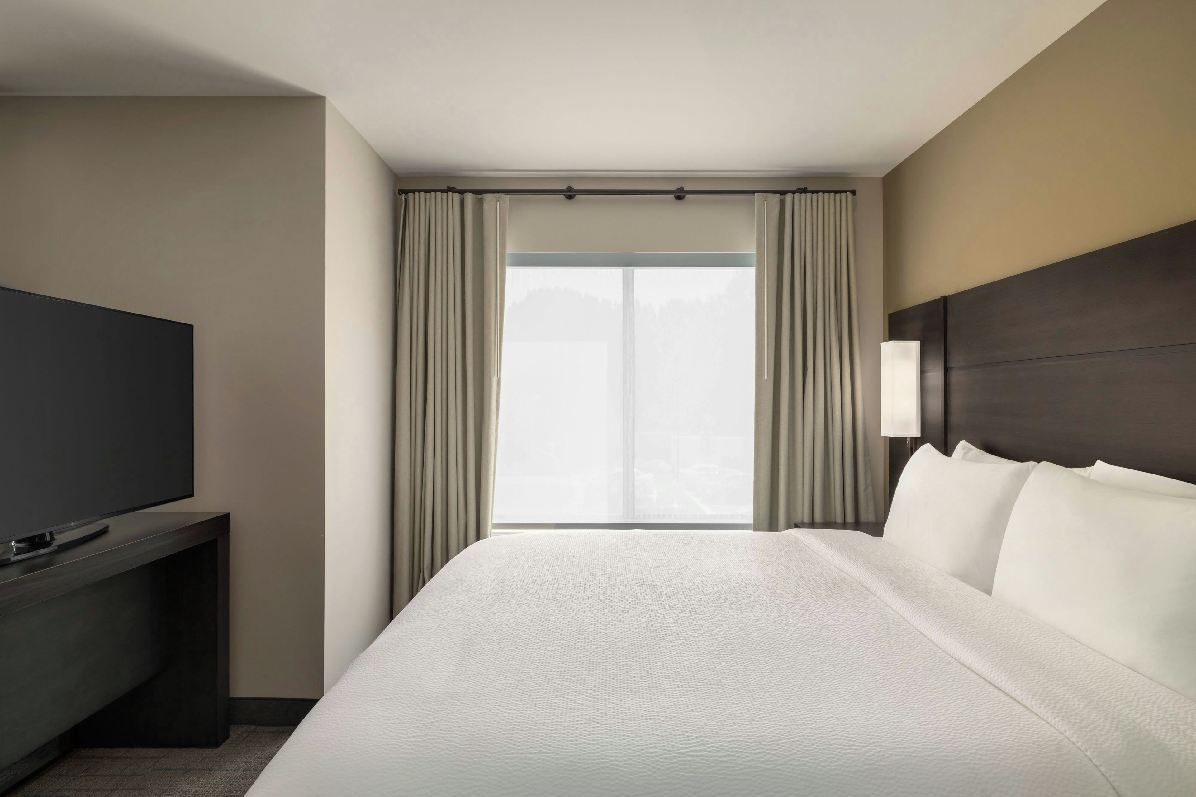A hotel room with a bed, a TV on a stand, a window with curtains, and a wall-mounted light.