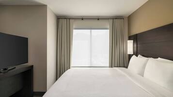 A hotel room with a bed, a TV on a stand, a window with curtains, and a wall-mounted light.
