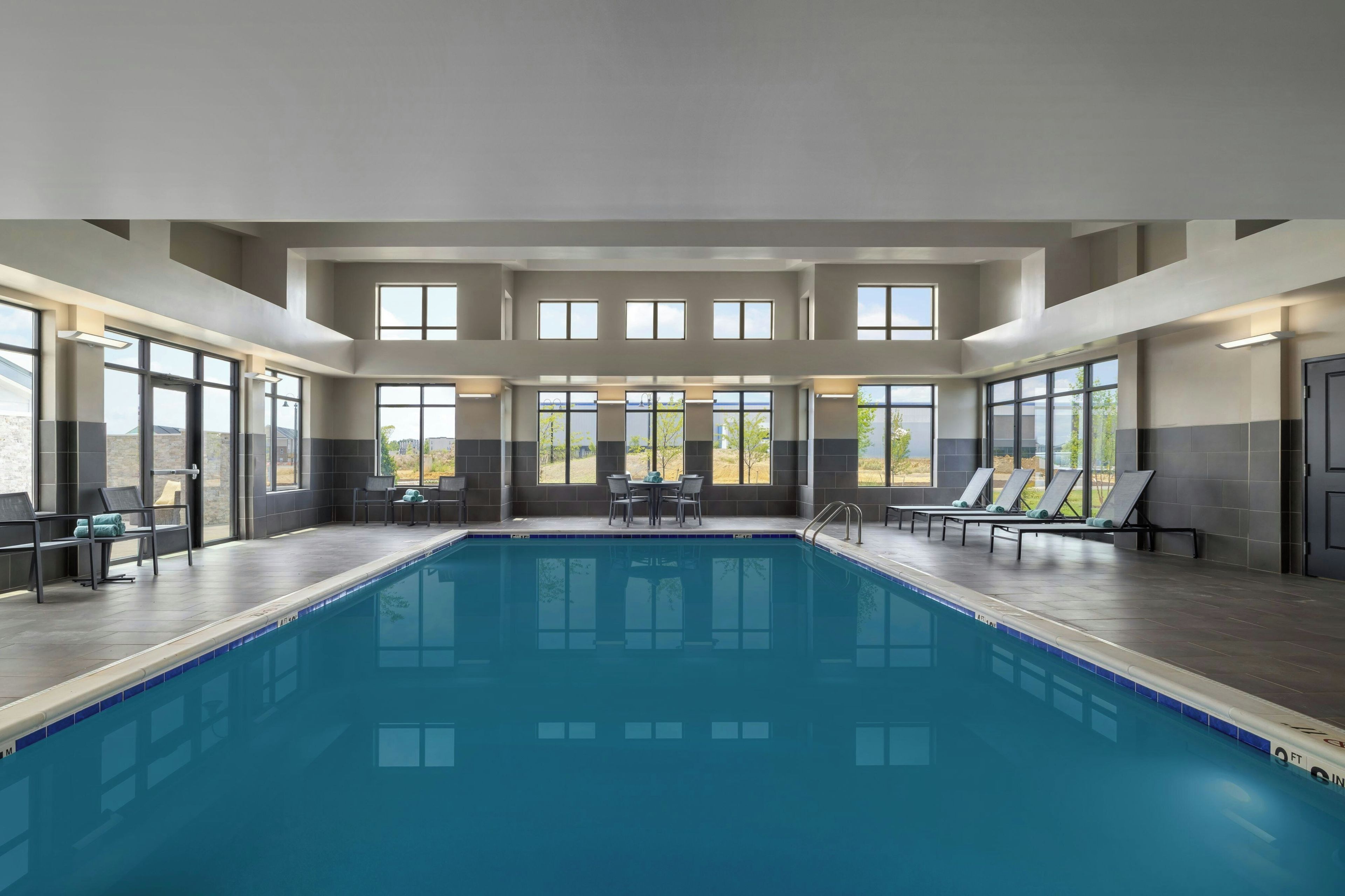 A large indoor swimming pool with multiple windows, lounge chairs, and a table with chairs.
