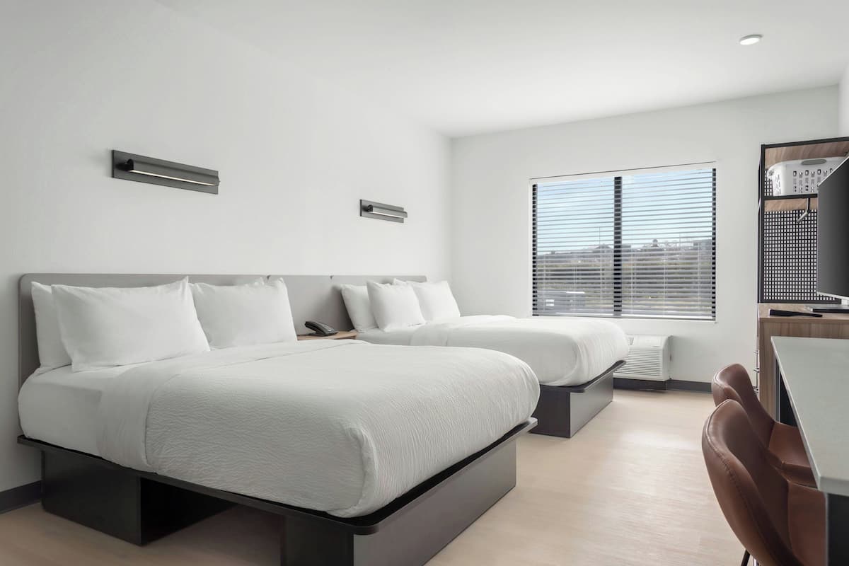 Una habitación de hotel moderna con dos camas, un escritorio y vista a la ciudad a través de amplios ventanales.