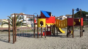 Un’area giochi con uno scivolo e strutture per arrampicata, circondata da una zona sabbiosa con palme e edifici sullo sfondo.