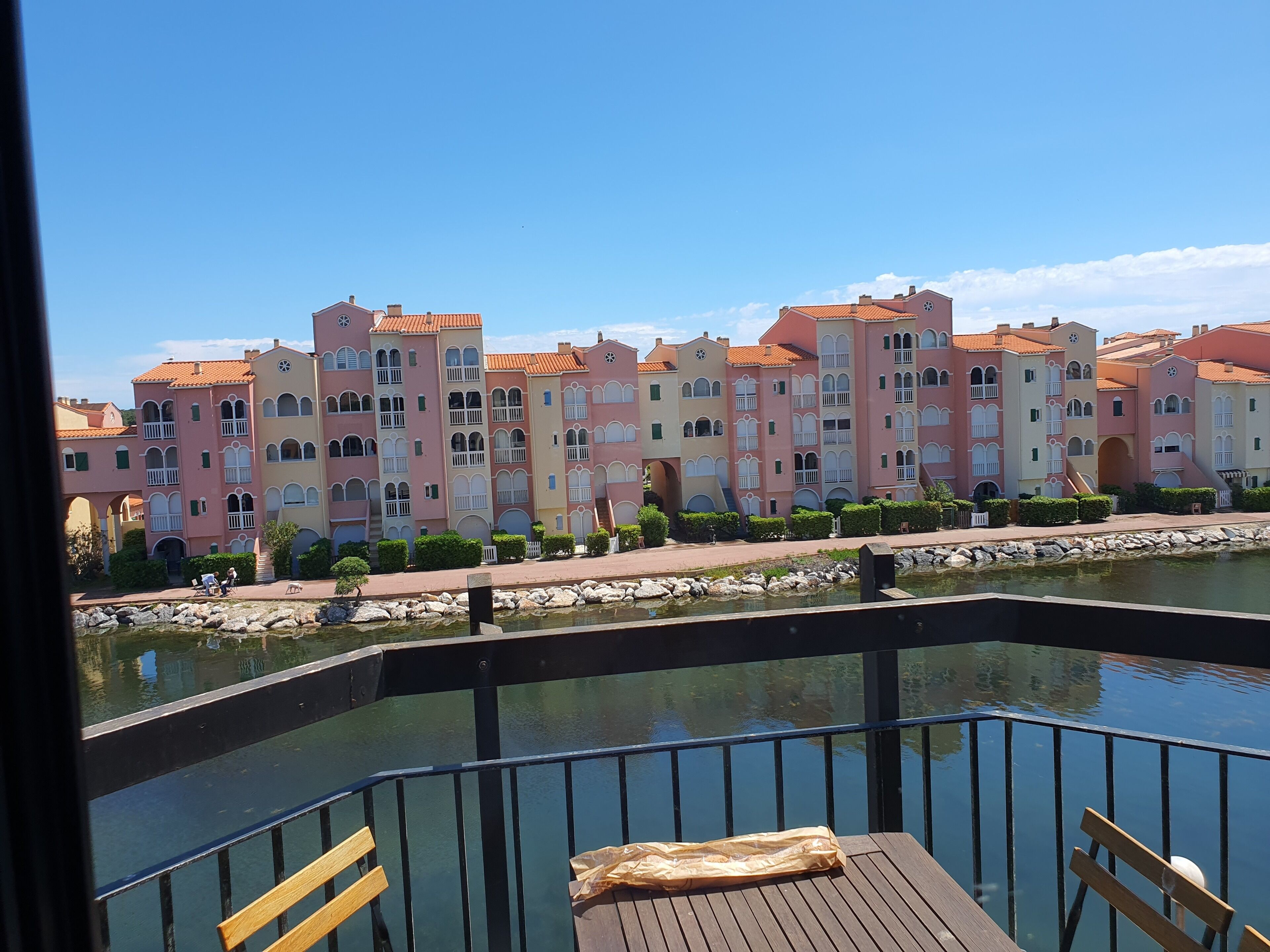 Vista para a orla marítima com uma fileira de edifícios coloridos, um deck de madeira com um banco e uma superfície aquática calma.