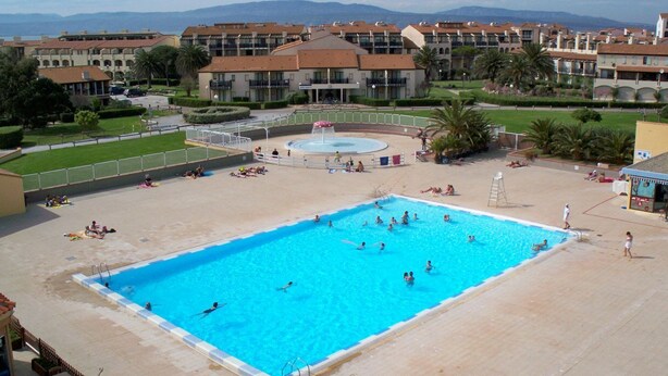 Una grande piscina circondata da un'area verde con palme e vista su una zona residenziale sullo sfondo.