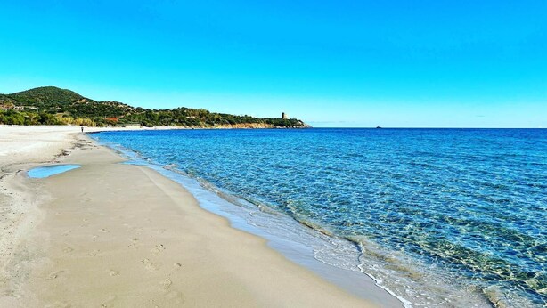 Uma praia com águas azul-claras, areal arenoso e uma colina com uma construção ao longe.