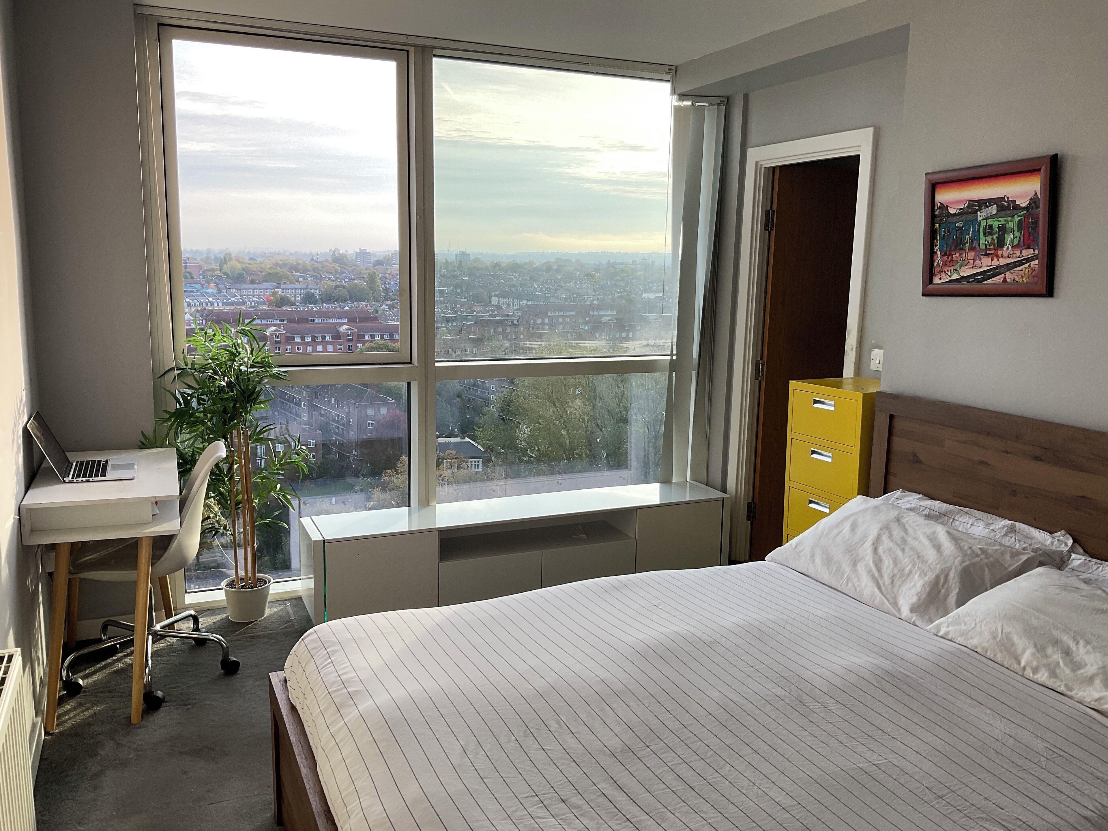 A bedroom with a large window, a desk with a laptop, a bed, a yellow dresser, and a potted plant.
