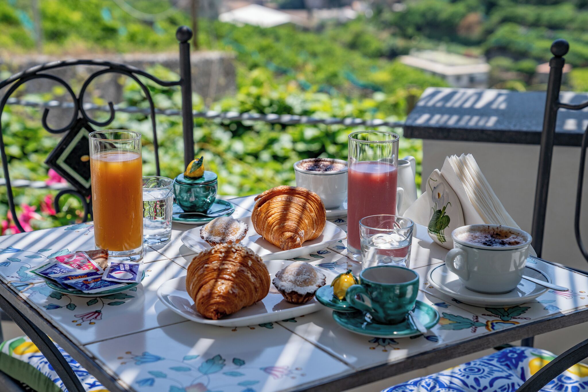 Una mesa servida con pasteles, bebidas y una taza de café en un balcón con vista a un jardín.