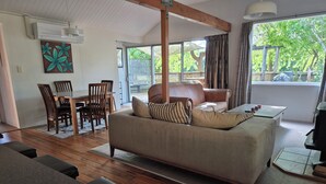 A cozy living room with a sofa, dining table, and chairs. There is a large window with a view of greenery outside.