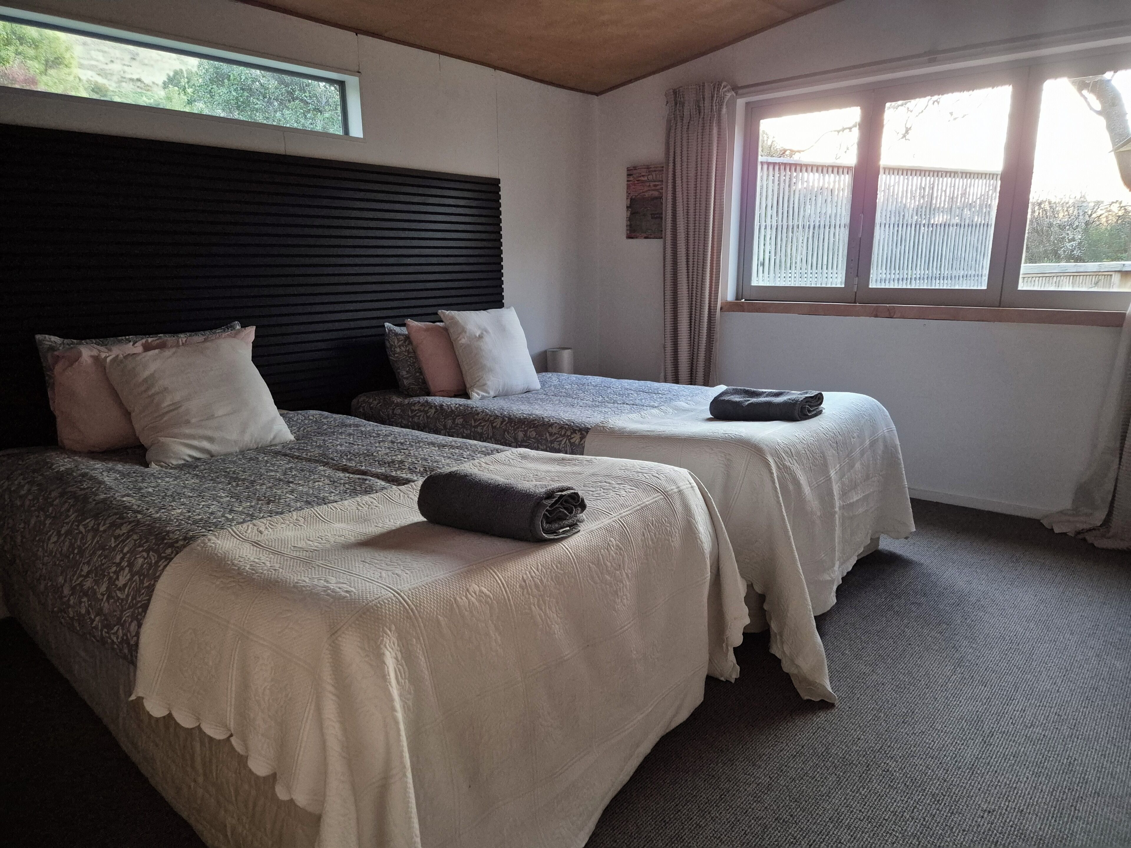A room with two beds, each with a headboard, a window with curtains, and a view of the outside.