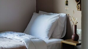Ein ordentlich bezogenes Bett mit weißen Leinen, ein an der Wand hängendes abstraktes Gemälde, ein Nachttisch mit Lampe und eine Vase mit getrockneten Blumen.