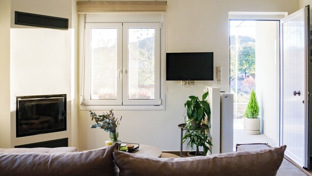 Ein Wohnzimmer mit einer Couch, einem Wandfernseher, einem Fenster mit Jalousien und einer Tür, die zu einem Außenbereich führt.