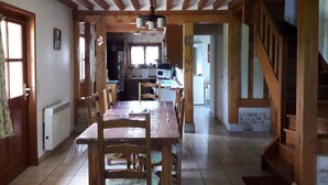 A spacious dining area with a wooden table and chairs, a kitchen in the background, and a staircase leading to an upper level. - Farm cottage in a quiet location near the sea (Beaufour-Druval)