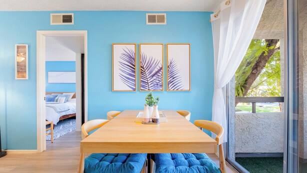 A room with a wooden dining table, blue cushioned chairs, a bed with white bedding, and a large window with a view of greenery. - Designer Resort Style - 1 King Bed Apartment, Pool, Gym in beautiful Gilbert AZ (Gilbert)