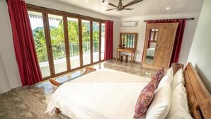 A spacious bedroom with a large bed, a wooden desk, and a view of greenery through large windows. - LA BELLE RESIDENCE (Mahe Island)