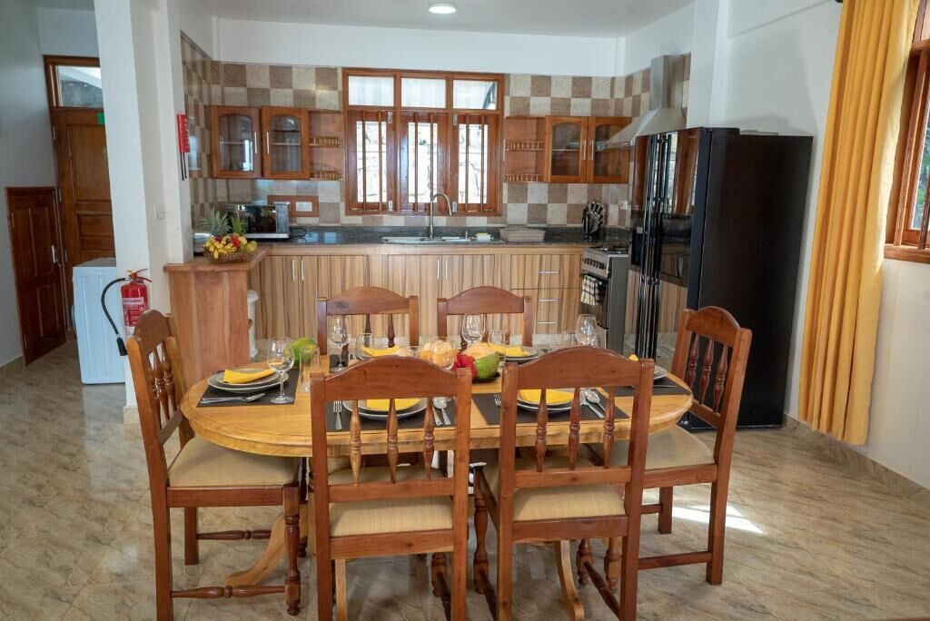 A dining area with a wooden table and chairs, a kitchen with checkered tiles and wooden cabinets, and a black refrigerator.
