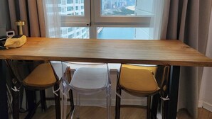A modern room with a wooden table and two chairs, a large window with curtains, and a view of a cityscape. - Seaview & City Lights Retreat, Marine City (Busan)