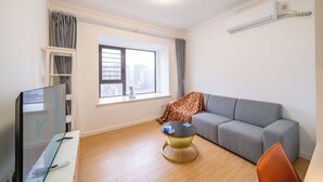 Una sala moderna con un sofá gris, una mesita redonda con un libro y un bolígrafo, un televisor sobre un mueble bajo y una ventana con cortinas.