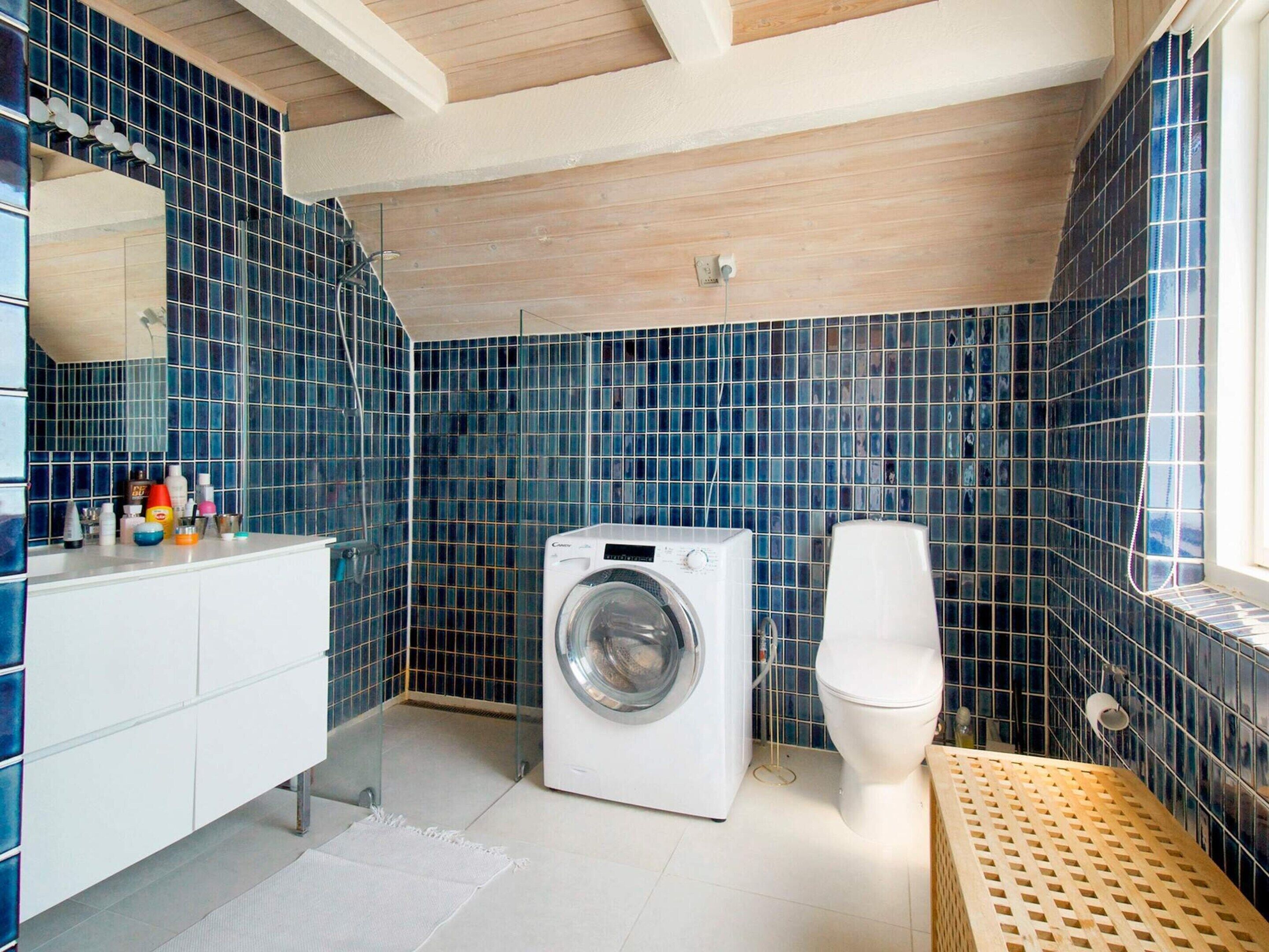 A compact bathroom with blue-tiled walls, a washing machine, a toilet, and a wooden bench.