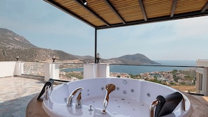 Eine große, runde Whirlwanne mit mehreren Wasserhähnen, auf einem Balkon mit Blick auf eine gebirgige Landschaft und das Meer.