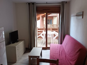 A small room with a sofa, a table, a TV on a cabinet, and a sliding glass door leading to an outdoor area with a wooden deck and chairs. - Ski Apartment With Pool & Free Parking (Les Angles)