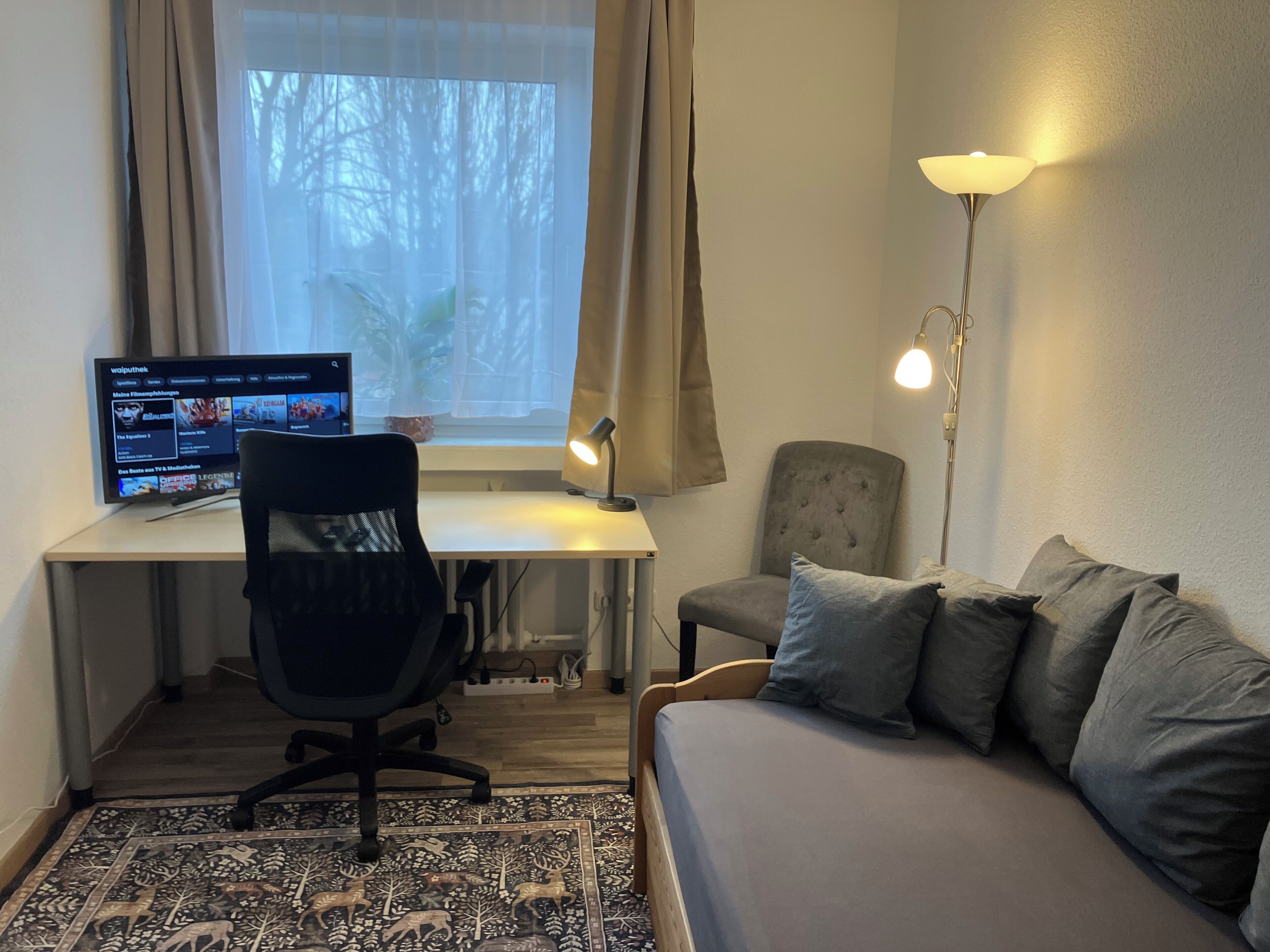 A room with a desk, a computer, a chair, a sofa, and a floor lamp.
