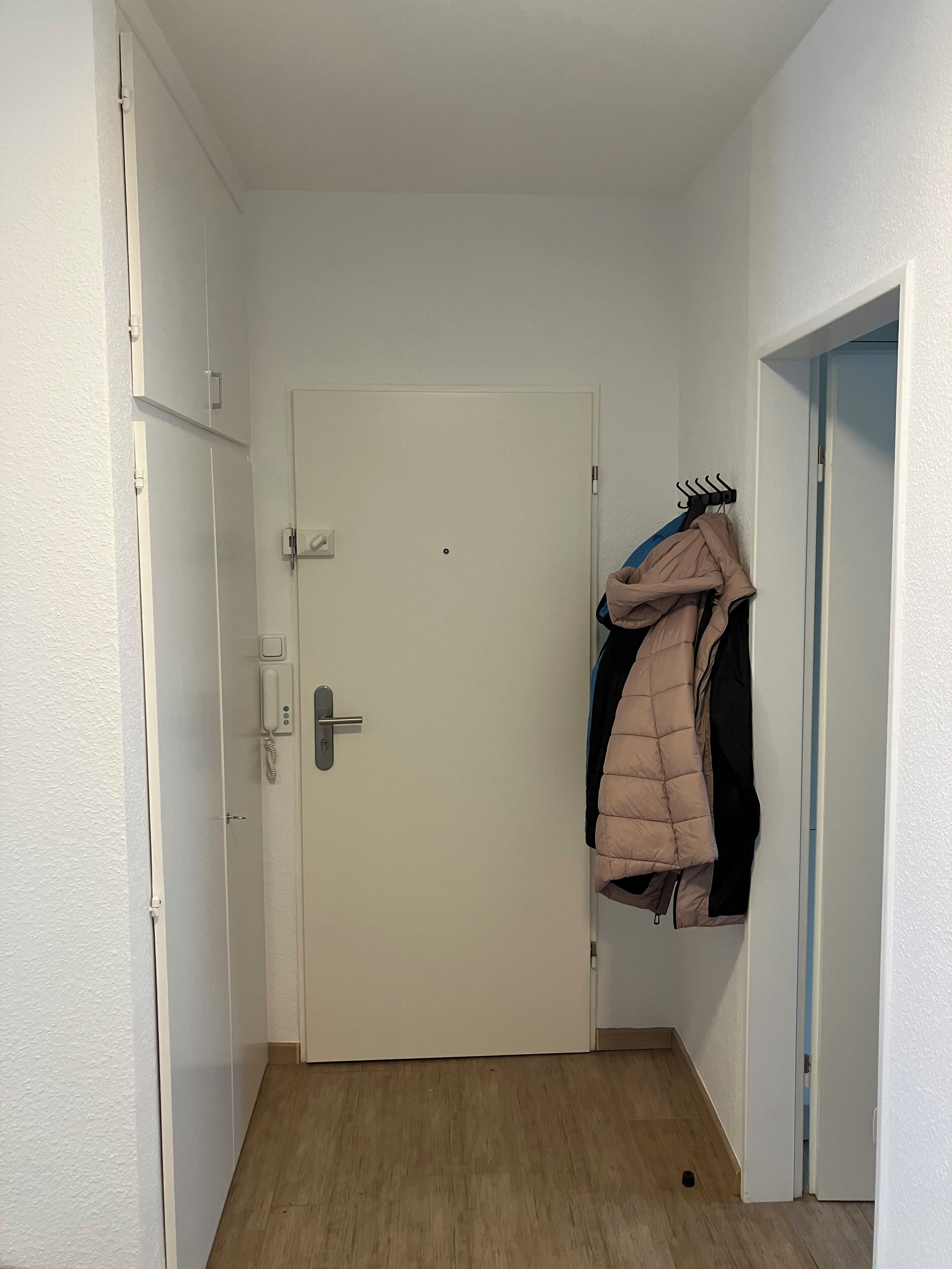 Un pasillo con una puerta blanca, un perchero con una chaqueta color beige y un teléfono en la pared.