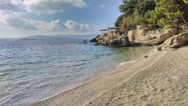 A pebble beach with clear water, rocky cliffs, and sparse vegetation.