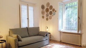 A living room with a grey sofa, a small side table, a radiator, and a window with a view of greenery. - 60sqm downtown with garden | Casa Da Diego (Civitanova Marche)